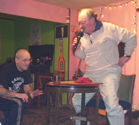 Gordon Cragg standing up besides bingo table and reaching into his back pocket while Oz checks the alignment of the glass on the table prior to trying to throw bits of paper in it